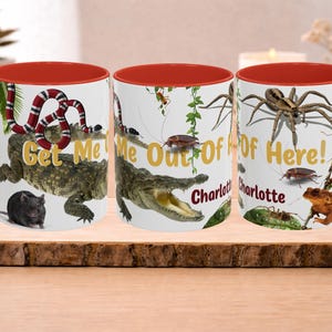 Può includere: Tre tazze in ceramica bianca con interni e manici rossi. Ogni tazza presenta un design diverso: un serpente e un alligatore, un alligatore e insetti, e un ragno e insetti. Le tazze hanno il testo "Get Me Out Of Here!" e un nome.