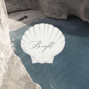 Op de afbeelding: Een witte sint-jakobsschelpafbeelding in het midden, met het woord Bright in een zwart cursief handschrift op het oppervlak. De schelp is geplaatst over een uitzicht op een zandstrandbaai met lichtblauw water en rotsachtige kliffen.