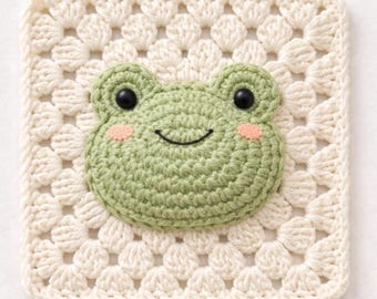 May include: A square, cream-colored crocheted item featuring a green frog face. The frog has black eyes and pink cheeks. The background is a granny square pattern.