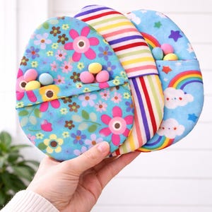 Könnte beinhalten: Drei eiförmige Stoffbeutel. Einer ist hellblau mit rosa Blumen, ein anderer hat Regenbogenstreifen und der dritte ist hellblau mit einem Regenbogen und Wolken. Jeder Beutel enthält kleine, bunte Süßigkeiten.