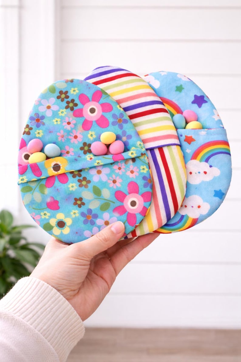 Op de afbeelding: Drie eivormige stoffen zakjes. E&eacute;n is lichtblauw met roze bloemen, een andere heeft regenboogstrepen en de derde is lichtblauw met een regenboog en wolken. Elk zakje bevat kleine, kleurrijke snoepjes.