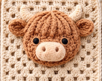 May include: A crocheted square featuring a Highland cow face. The cow has brown fur, black eyes, and cream-colored horns and muzzle. The square is made of cream-colored yarn with a granny square pattern.