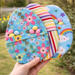 Op de afbeelding: Een hand houdt drie eivormige stoffen zakjes voor Pasen vast. De zakjes hebben bloemen-, regenboog- en sterrenpatronen. Elk zakje bevat kleurrijke snoepjes. De zakjes zijn in tinten blauw, roze, geel en rood.