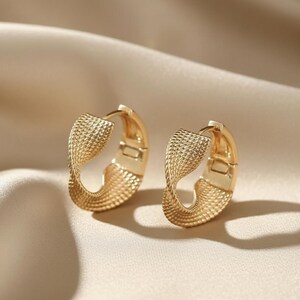 May include: A pair of gold-coloured hoop earrings. The earrings feature a textured, woven design and a hinged closure. They are presented against a neutral-toned background.