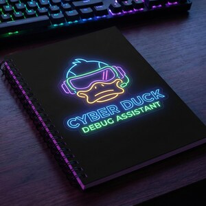 May include: A black notebook with the text "CYBER DUCK DEBUG ASSISTANT" in neon green and blue, and a neon duck wearing VR goggles. A backlit keyboard and mouse are visible in the background, on a wooden surface.