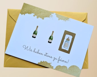 Pode incluir: Um cartão azul claro com detalhes dourados e o texto "Wir haben etwas zu feiern!". Apresenta ilustrações de garrafas de champanhe e uma mamadeira. O cartão está dentro de um envelope dourado.