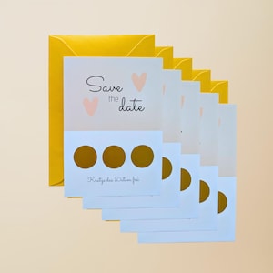 May include: A stack of white "Save the Date" cards with gold scratch-off circles and pink heart accents, accompanied by a yellow envelope. The cards have the text "Save the date" and "Kratze das Datum frei".