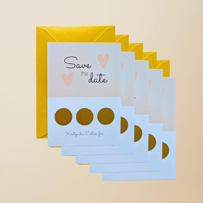 May include: A stack of white "Save the Date" cards with gold scratch-off circles and pink heart accents, accompanied by a yellow envelope. The cards have the text "Save the date" and "Kratze das Datum frei".