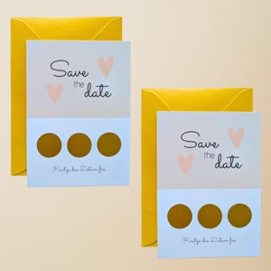 May include: Two "Save the Date" cards with yellow envelopes. The cards have a light blue section with three gold scratch-off circles. Above, the cards read "Save the date" with two pink hearts. The bottom of the card reads "Kratze das Datum frei".