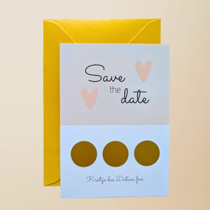 May include: A light blue and white "Save the Date" card with two pink hearts and the text "Save the date". The card has three gold scratch-off circles and the text "Kratze das Datum frei". A yellow envelope is behind the card.