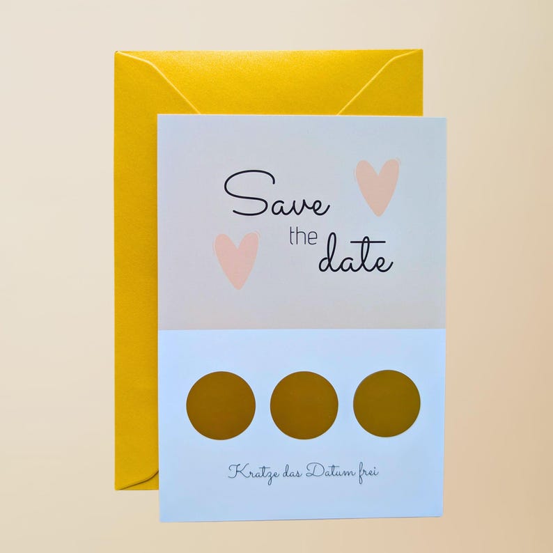 May include: A light blue and white "Save the Date" card with two pink hearts and the text "Save the date". The card has three gold scratch-off circles and the text "Kratze das Datum frei". A yellow envelope is behind the card.