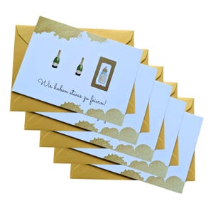 May include: A stack of greeting cards with gold envelopes. The cards have a light blue background with gold accents and illustrations of champagne bottles and a baby bottle. The text on the card reads "Wir haben etwas zu feiern!"