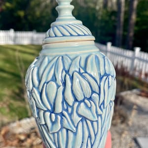 May include: A light blue ceramic jar with a lid. The jar is decorated with a floral pattern and has a rounded shape. The lid has a pointed finial. The jar is held in a person's hand.