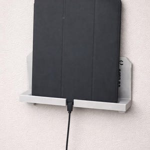 May include: A black tablet with a folio case rests on a white shelf attached to a textured white wall. A black charging cable extends from the tablet. The shelf is a simple, minimalist design.
