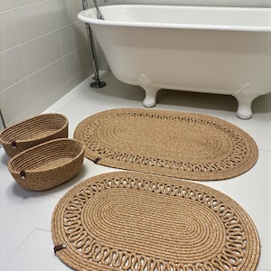 Op de afbeelding: Een badkamer met twee ovale jute badmatten en twee bijpassende geweven manden. De matten en manden hebben een natuurlijke bruine kleur. Op de achtergrond staat een witte badkuip met leeuwenpoten.