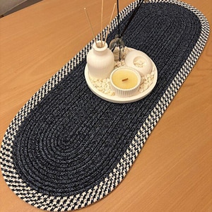 May include: An oval-shaped table runner crafted from dark blue braided rope with a white and blue chequered border. Centred on the runner is a round white tray containing a white ribbed vase with reed diffusers and a small yellow candle.