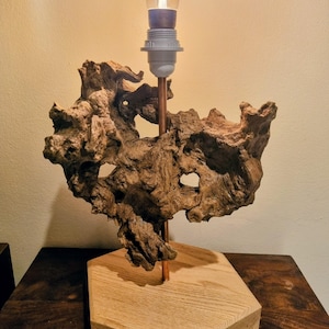 Op de afbeelding: Een unieke tafellamp met een verweerde, bruine drijfhoutsculptuur. De lamp heeft een koperen staaf en een gloeilamp met een zichtbare gloeidraad. De basis is een lichtgekleurd, zeshoekig houten blok.