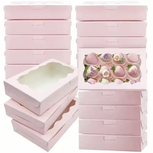 May include: A collection of light pink bakery boxes, some with clear window cutouts. One box displays chocolate-covered strawberries decorated with flowers. The boxes are stacked, suggesting they are for packaging treats.