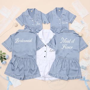 May include: A collection of five short-sleeved pajama sets in dusty blue and white satin. The blue sets feature white piping and embroidered names like Bea, Gabriella, Lena, Bridesmaid, and Maid of Honor. The white set has black piping.