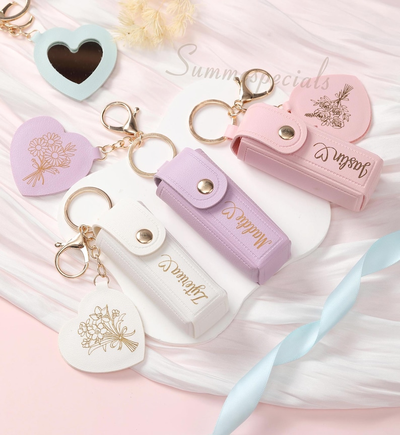 May include: A collection of keychains in pastel colors, including heart-shaped mirrors and cases. Some keychains feature floral designs and personalized names. Gold-colored hardware and clasps are used throughout the collection. The text "Summer Specials" is visible in the background.