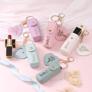 May include: A collection of personalized lipstick holders in pastel colors, with gold keychains and heart-shaped charms. Each holder is engraved with a name. A red lipstick is also visible.