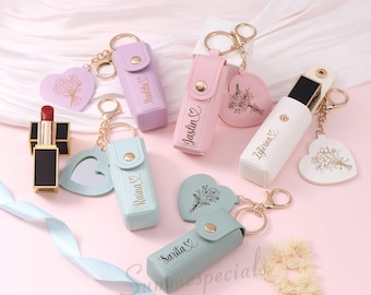 May include: A collection of personalized lipstick holders in pastel colors, with gold keychains and heart-shaped charms. Each holder is engraved with a name. A red lipstick is also visible.