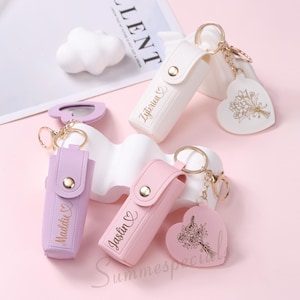 May include: A collection of personalized keychains in white, lavender, and pink. Each keychain features a rectangular case with a snap closure and a heart-shaped charm. The names "Zyterica", "Maddie", and "Jaslin" are printed on the cases.