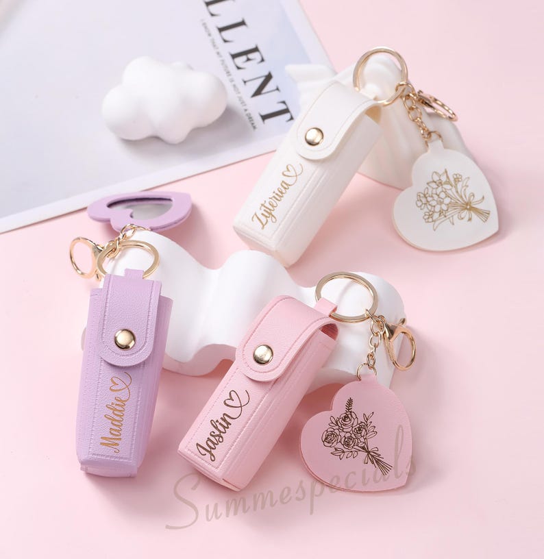 May include: A collection of personalized keychains in white, lavender, and pink. Each keychain features a rectangular case with a snap closure and a heart-shaped charm. The names "Zyterica", "Maddie", and "Jaslin" are printed on the cases.