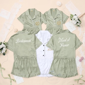 May include: A set of sage green and white pyjama sets with short sleeves and shorts. The sage green tops feature the text Bridesmaid, Maid of Honor, and gold embroidered initials. The white top features the gold embroidered text Bride.