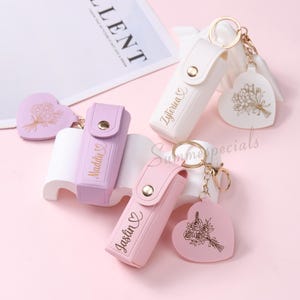 May include: A collection of personalized keychains in white, lavender, and pink. Each keychain features a holder with a name and a heart-shaped charm with a floral design. Gold-colored hardware connects the holder and charm.