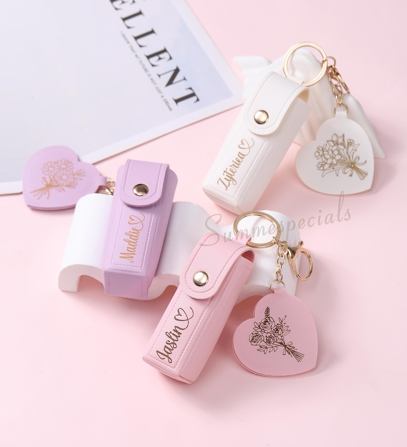 May include: A collection of personalized keychains in white, lavender, and pink. Each keychain features a holder with a name and a heart-shaped charm with a floral design. Gold-colored hardware connects the holder and charm.