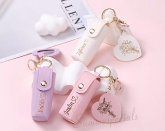 May include: A collection of personalized keychains in white, lavender, and pink. Each keychain features a small case with a gold button clasp and a heart-shaped charm with a floral design. The names "Zyterica", "Maddie", and "Jaslin" are printed on the cases.