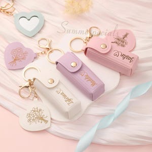May include: A collection of keychains in pastel colors, including heart-shaped mirrors and cases. Some keychains feature floral designs and names in gold lettering. The keychains are attached to gold-colored metal rings and clasps.