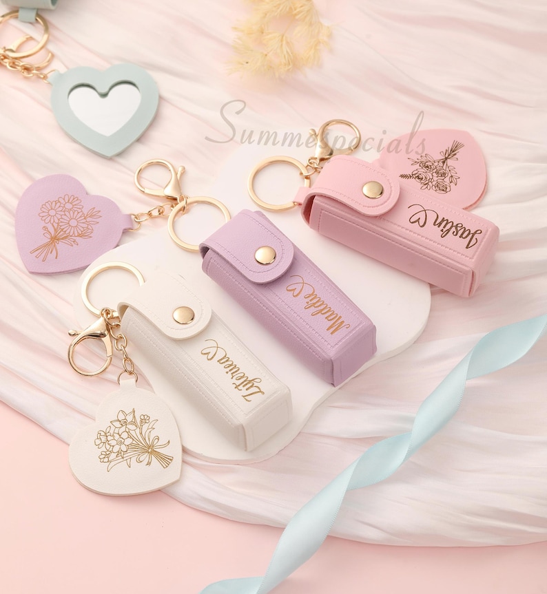 May include: A collection of keychains in pastel colors, including heart-shaped mirrors and cases. Some keychains feature floral designs and names in gold lettering. The keychains are attached to gold-colored metal rings and clasps.
