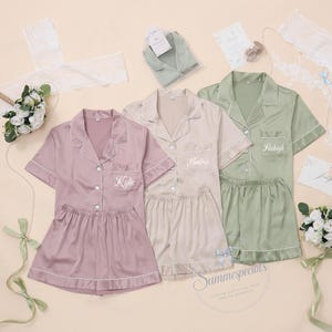 Op de afbeelding: Drie sets van pyjamashirts met korte mouwen en knoopsluiting en bijpassende shorts in mauve, champagne en saliegroen satijn. Elk shirt heeft een borstzak die is geborduurd met de namen Kylie, Andrea en Raleigh in wit schrift.