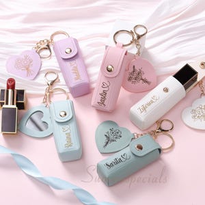 May include: A collection of personalized lipstick holders and keychains in various pastel colors, including pink, lavender, and blue. Each holder has a snap closure and is engraved with a name. Heart-shaped keychains with floral designs and a lipstick are also present.