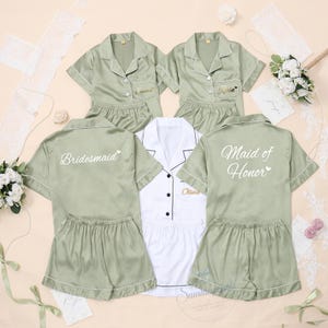 May include: A collection of five short-sleeved pyjama sets in sage green and white satin fabric. The tops feature button-down fronts and collars. The backs of the green shirts are printed with the words Bridesmaid and Maid of Honor in white.