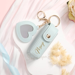 May include: A light blue keychain with a heart-shaped mirror and a matching case. The case has the name "Minji" in gold lettering and a gold button closure. The keychain is attached to a gold ring and chain.