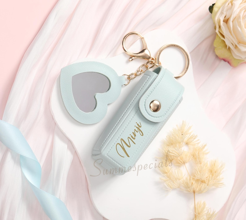 May include: A light blue keychain with a heart-shaped mirror and a matching case. The case has the name "Minji" in gold lettering and a gold button closure. The keychain is attached to a gold ring and chain.