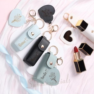 May include: An assortment of lipstick holders and lipsticks. The holders are in various colors, including black, light blue, and white, and feature gold keychains. Some holders have names printed on them. A red lipstick is also visible.
