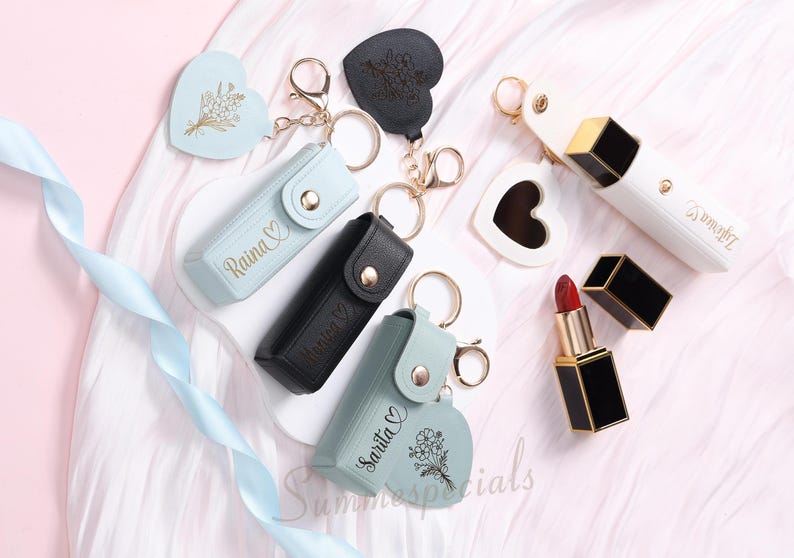 May include: An assortment of lipstick holders and lipsticks. The holders are in various colors, including black, light blue, and white, and feature gold keychains. Some holders have names printed on them. A red lipstick is also visible.