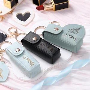 May include: A collection of lipstick holders in various colors, including black and light blue, with gold-colored keychains. Each holder has a snap closure and personalized text. Other items include lipsticks and a heart-shaped mirror.