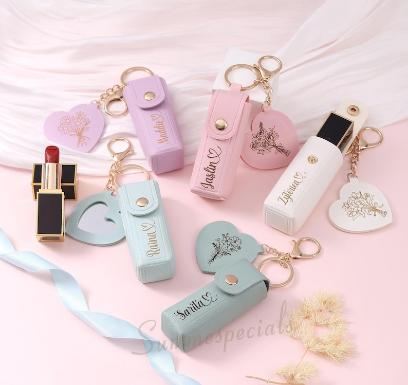 May include: A collection of personalized lipstick holders in pastel colors, with gold keychains and heart-shaped charms. Each holder is engraved with a name. A red lipstick is also visible.