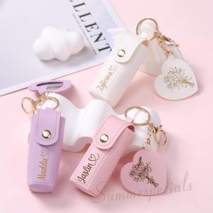 May include: A collection of personalized keychains in white, lavender, and pink. Each keychain features a small case with a gold button clasp and a heart-shaped charm with a floral design. The names "Zyterica", "Maddie", and "Jaslin" are printed on the cases.