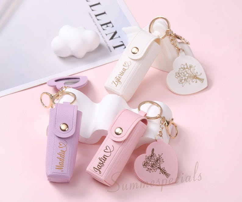 May include: A collection of personalized keychains in white, lavender, and pink. Each keychain features a small case with a gold button clasp and a heart-shaped charm with a floral design. The names "Zyterica", "Maddie", and "Jaslin" are printed on the cases.