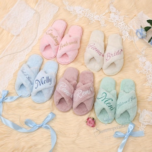 Op de afbeelding: Een collectie pluche slippers met open teen in pastelkleuren zoals roze, wit, lichtblauw, mauve en mintgroen. Elk paar is voorzien van geborduurde namen of titels zoals Lindsay, Bridesmaid, Fiona, Bride, Maid of Honor, Molly, Reni, Meni en Matron of Honor.