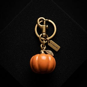 May include: A pumpkin-shaped charm keyring featuring a glossy orange pumpkin with a metallic stem and leaf. The charm is attached to a brass-toned metal split ring and a lobster clasp, accompanied by a small rectangular tag embossed with the word COACH.