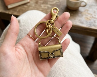 Könnte beinhalten: Ein messingfarbener Metallschlüsselanhänger mit einem Miniatur-Handtaschenanhänger, der einen Klappverschluss und ein markantes C-Logo aufweist. Der Anhänger ist an einem Ring und einem Karabinerhaken befestigt, inklusive eines kleinen Schilds mit COACH.