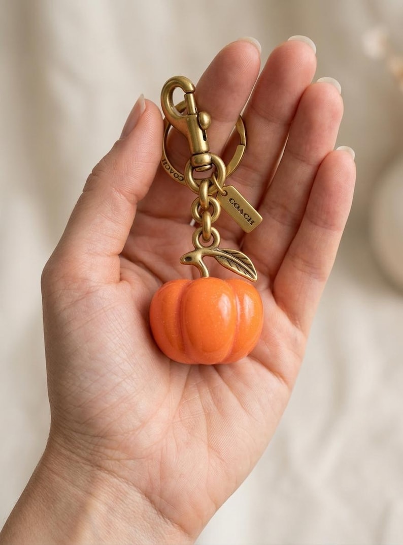 May include: A Coach brand keychain featuring a glossy orange pumpkin charm with a metallic stem and leaf. The charm is attached to a brass-toned lobster clasp and a split ring, with a small rectangular metal tag engraved with the Coach logo.