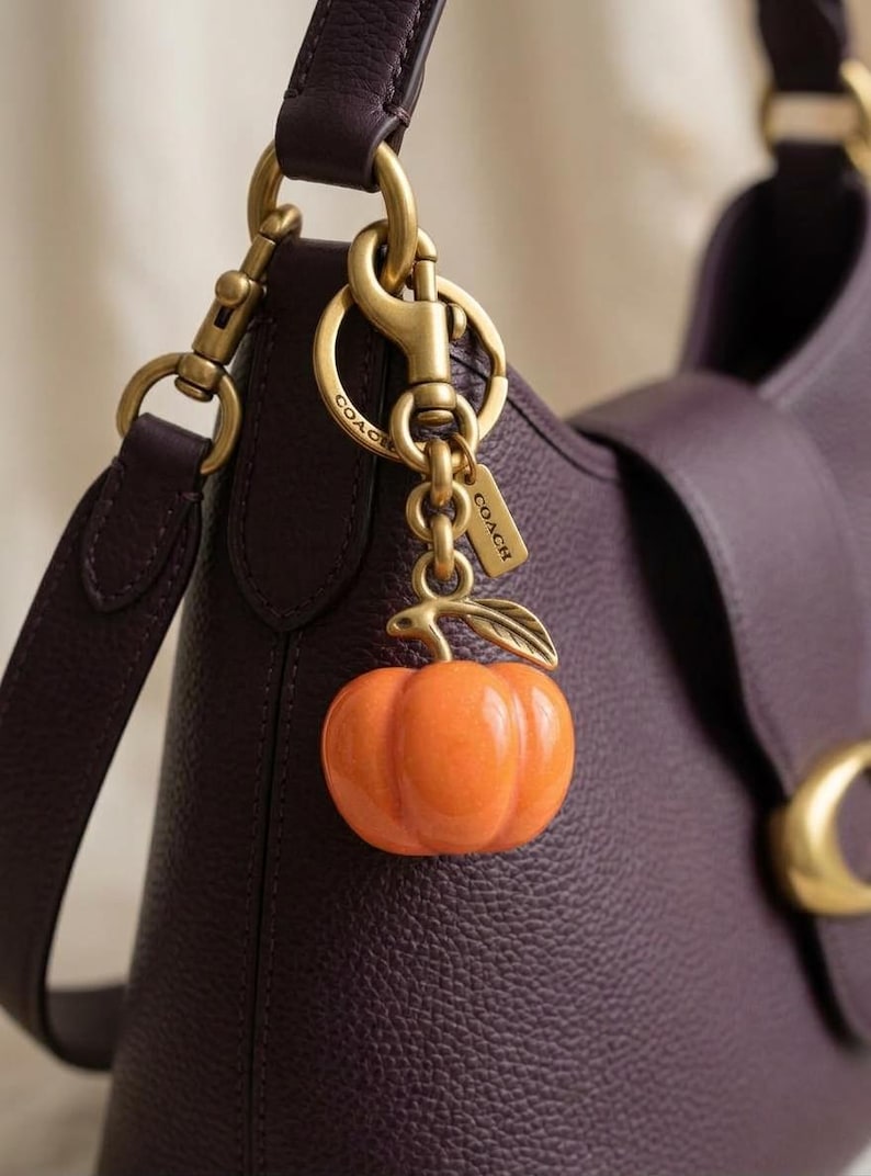 May include: A dark purple pebbled leather handbag featuring a gold-toned metal hardware clasp and a hanging bag charm. The charm consists of a glossy orange pumpkin pendant with a gold-toned leaf accent, attached to a gold-toned ring engraved with COACH.
